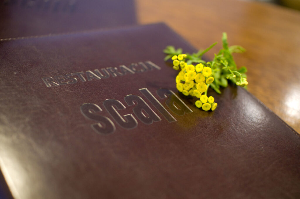 Bordowa skórzana okładka menu z tłoczonym napisem Restauracja Scala, obok żółte kwiaty wrotyczu