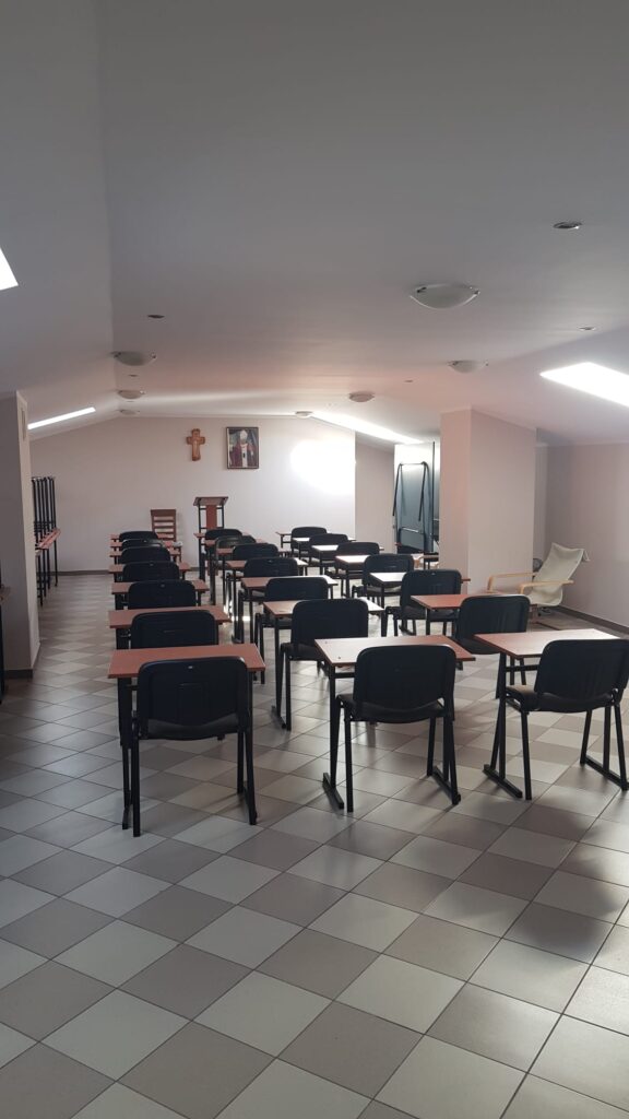 Rzędy stolików z czarnymi krzesłami zwrócone w stronę mównicy, na ścianie krzyż i portret