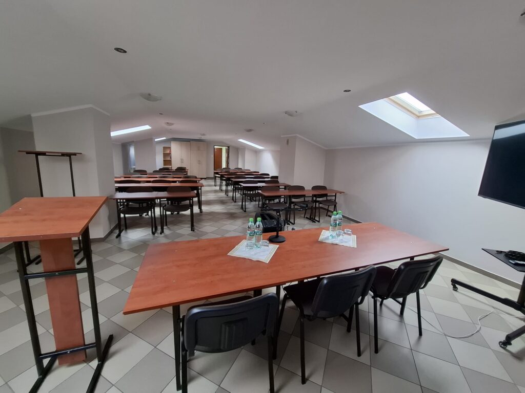Sala konferencyjna na poddaszu z oknami dachowymi, stół prezydialny, mównica i duży ekran