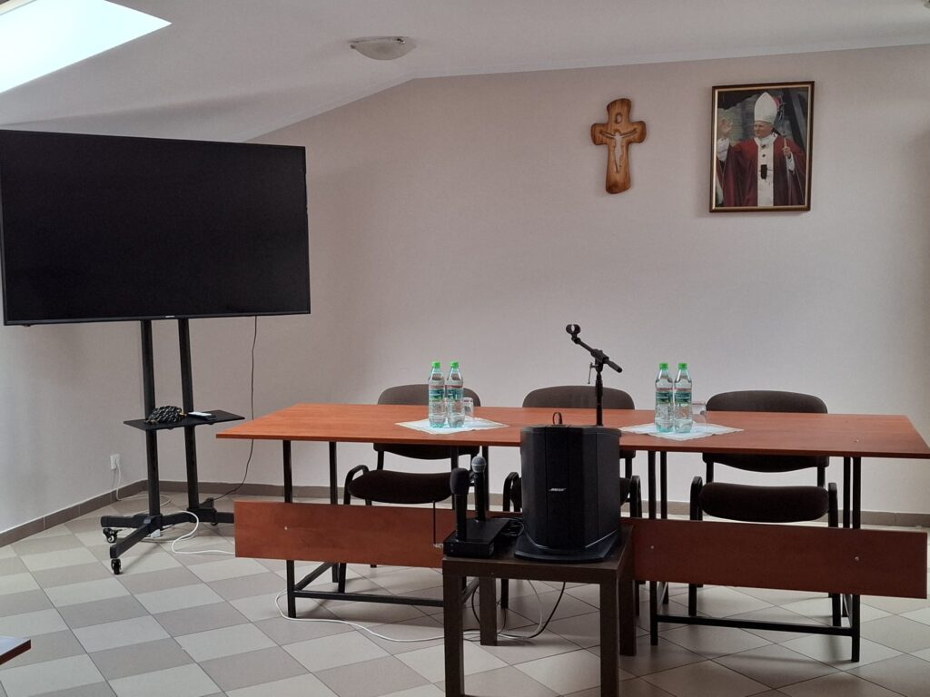 Stół prezydialny z mikrofonem i wodą, obok ekran na stojaku i głośnik, na ścianie krzyż i portret Jana Pawła II