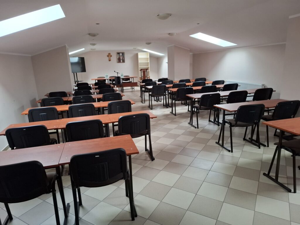 Sala na poddaszu z rzędami stolików i krzeseł, w głębi ekran, mównica i stół prezydialny